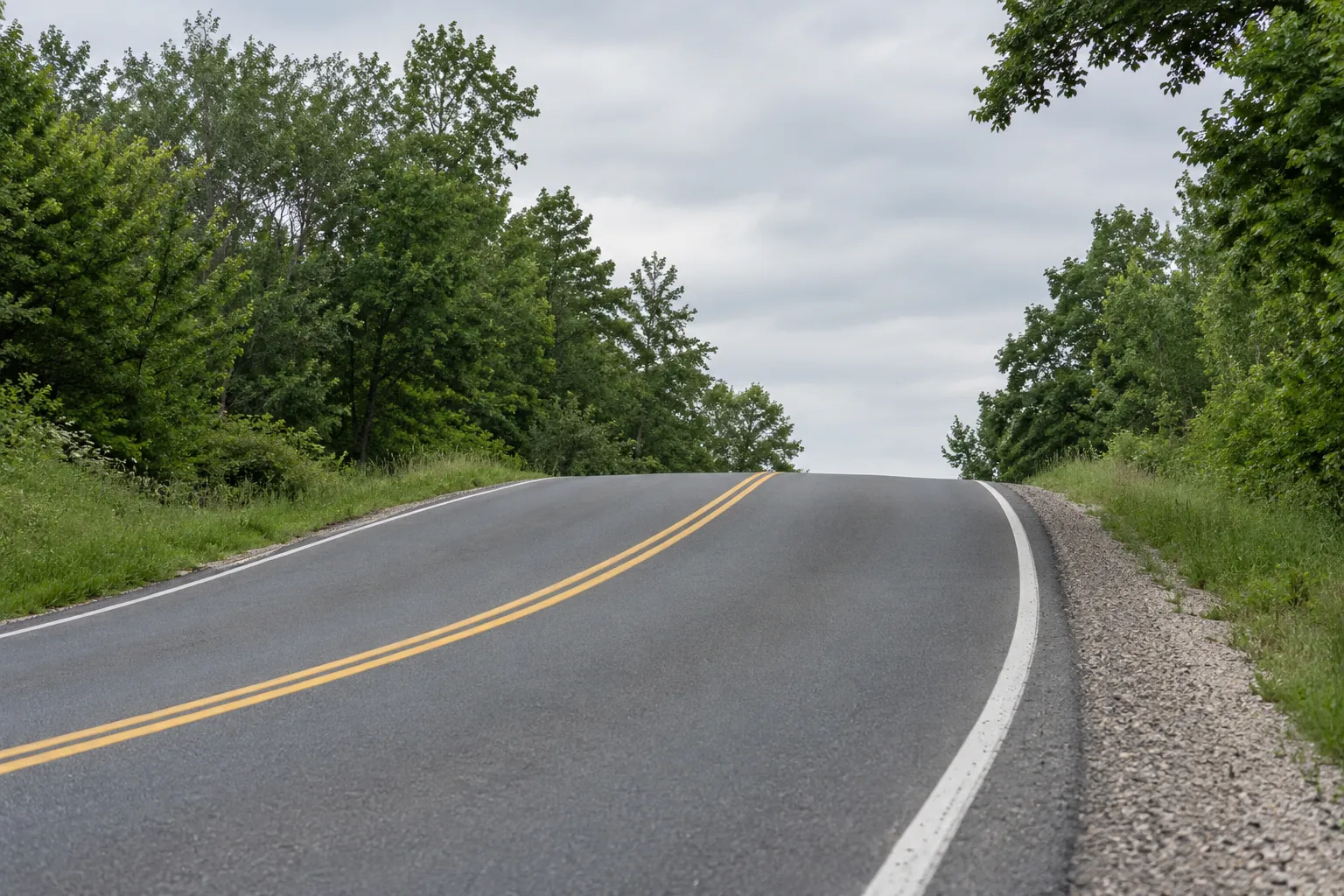 A two-lane rural road climbing over a hill crest and curving out of sight between trees.